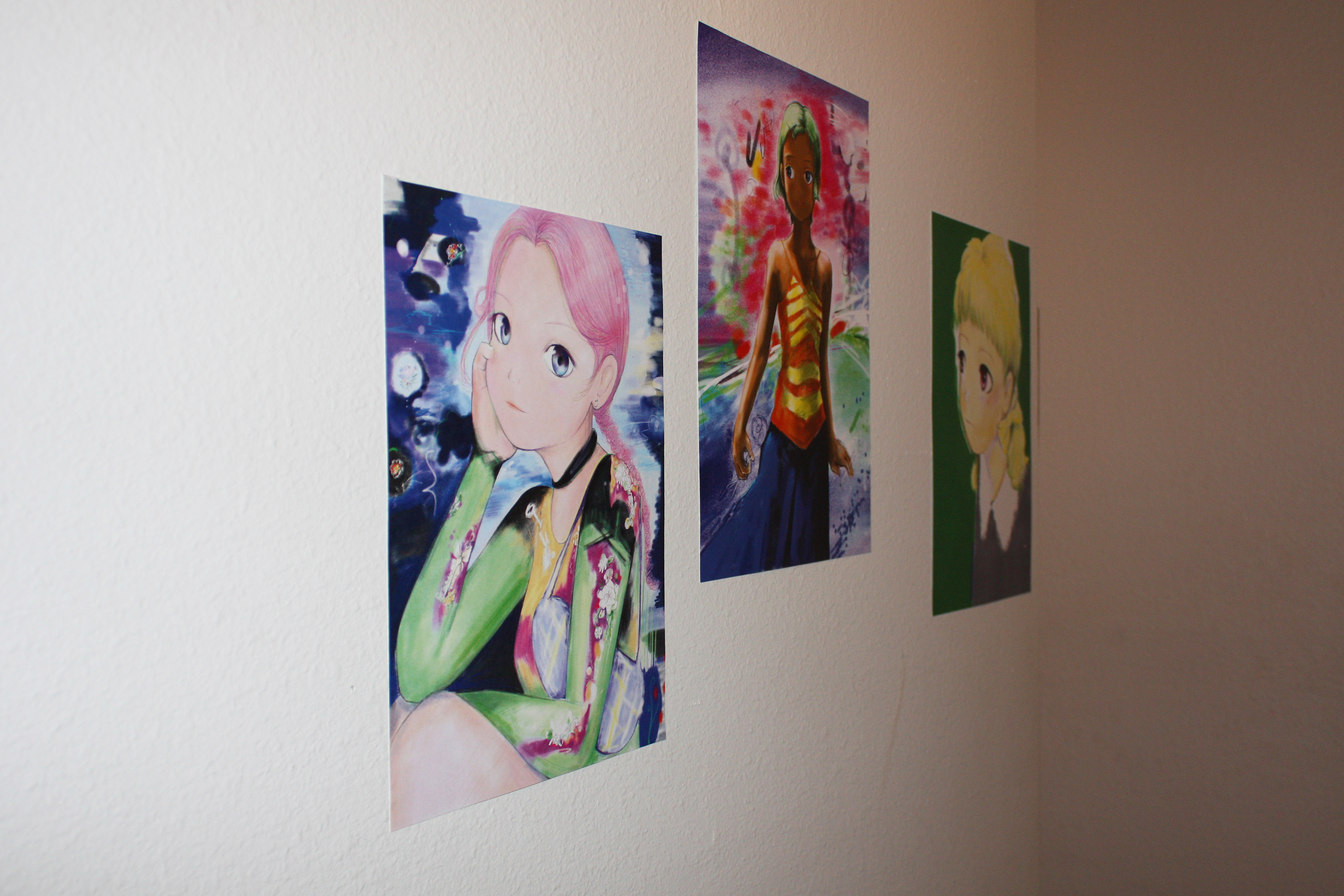 Three prints of anime style female figures hung in a narrow white hallway